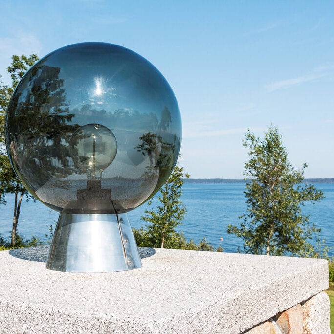 A glass orb with a lightbulb inside and a view of Penobscot Bay with the sun and trees reflecting off of the glass.