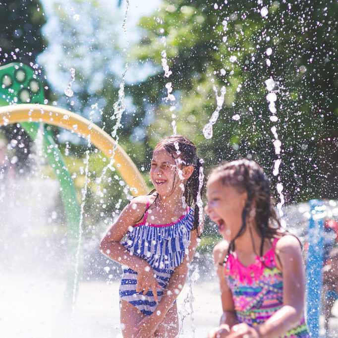 A group of young children frolic and run in a playground splash pad. It is a sunny, perfect day for getting wet and playing hard! Two young girls in swimsuits are running through the sprinklers and yelling and laughing as the water droplets cascade all around.